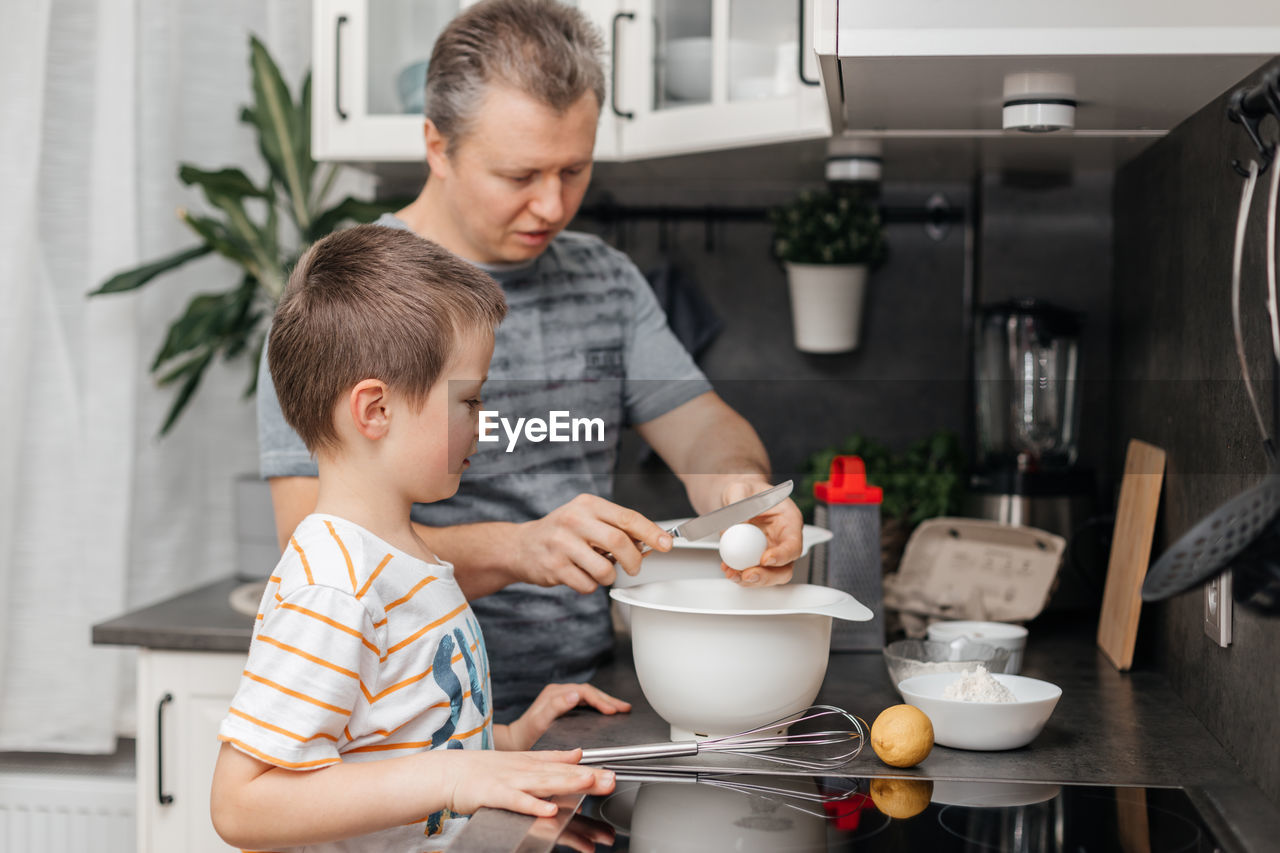 Dad and son are cooking in the kitchen. dad teaches to break the child's eggs and cook the dough.