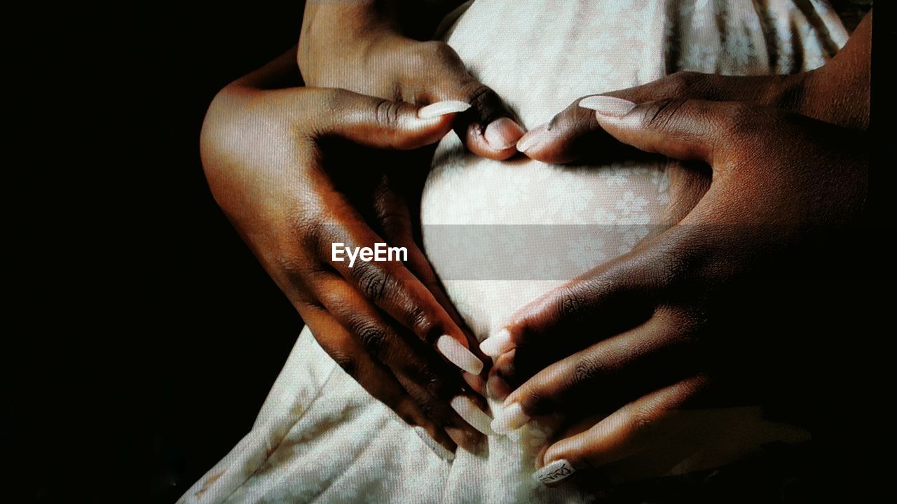 Close-up of couple hands making heart shape on pregnant abdomen against black background