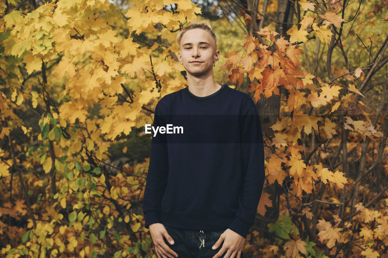 Confident teenage boy with blond hair standing against maple trees in autumn