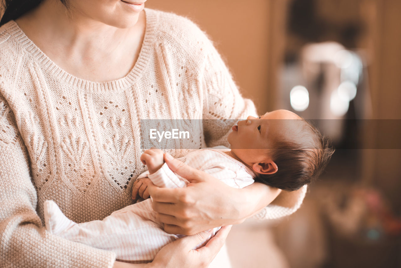 Smiling young woman holding cute little infant baby 1-2 months old on her hands in room at home