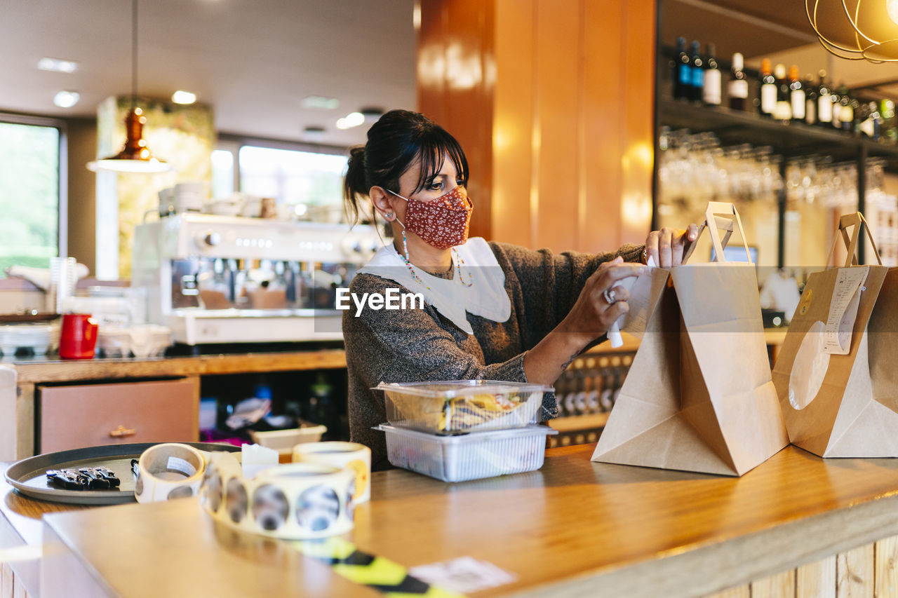 Female owner in mask attaching receipt on brown paper bag on counter at restaurant