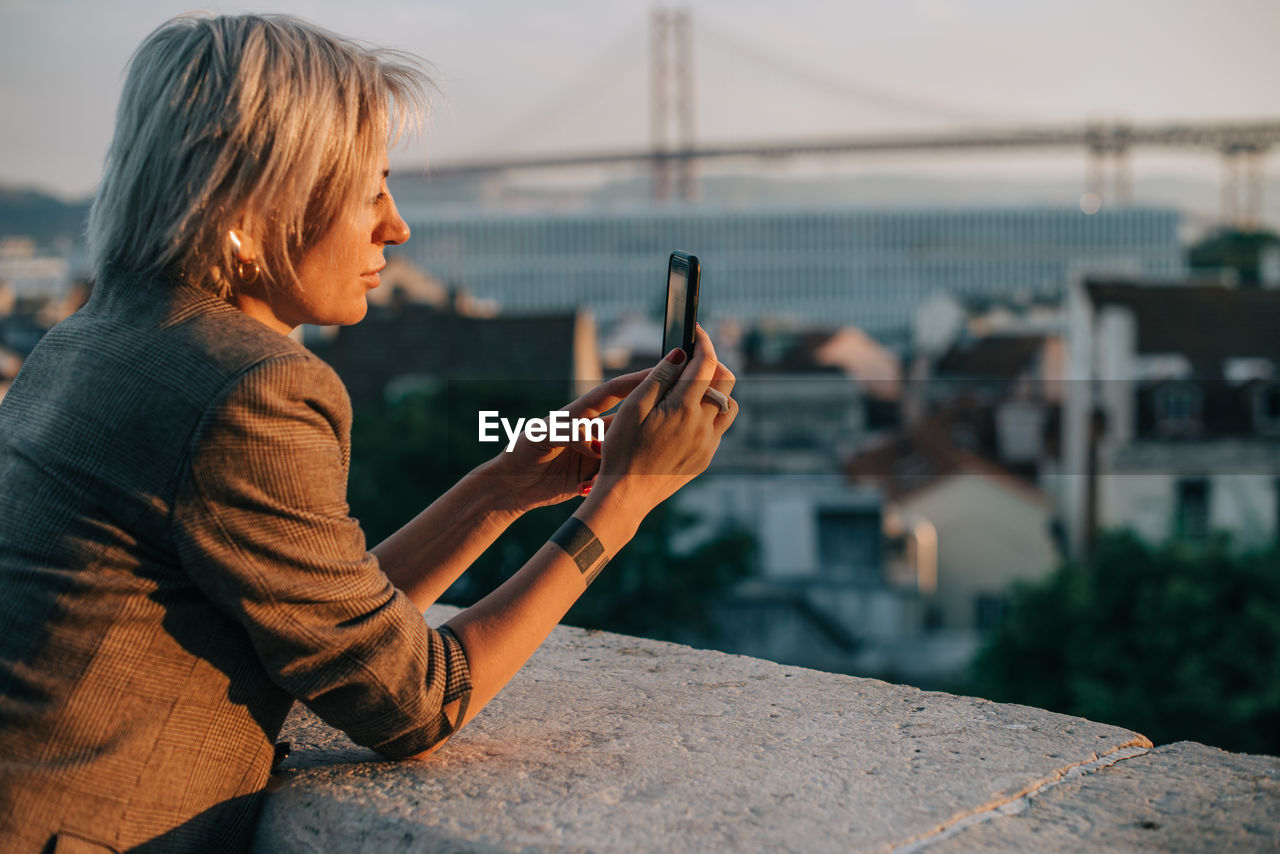 Blond woman in jacket taking cityscape photos with her smartphone