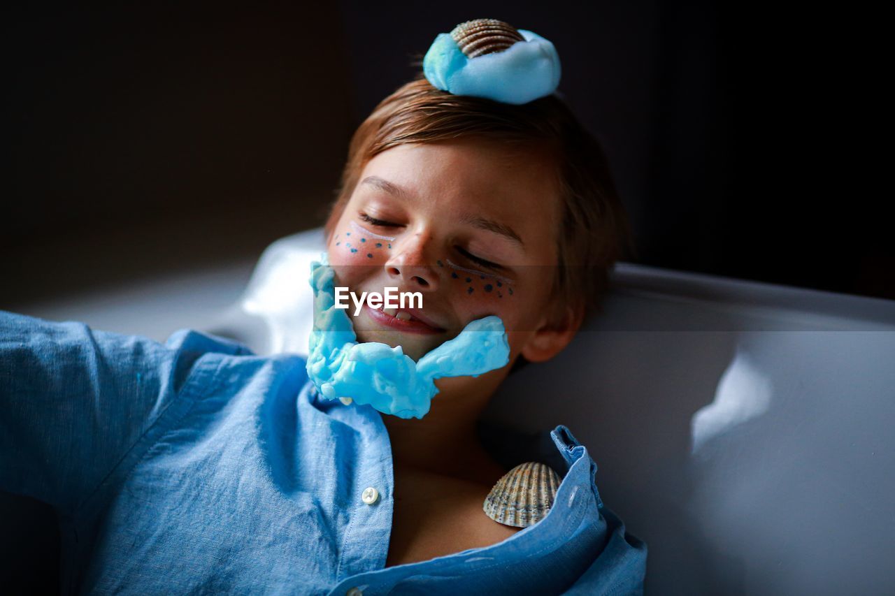 Close-up of boy with shaving foam and seashells on chair against black background