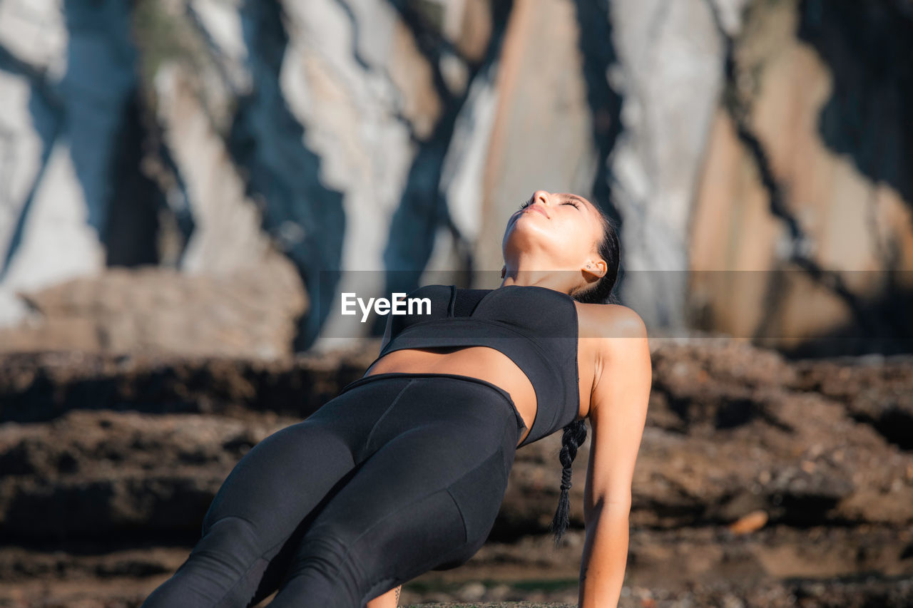 Calm female with closed eyes practicing yoga in purvottanasana on sunny day in rocky terrain