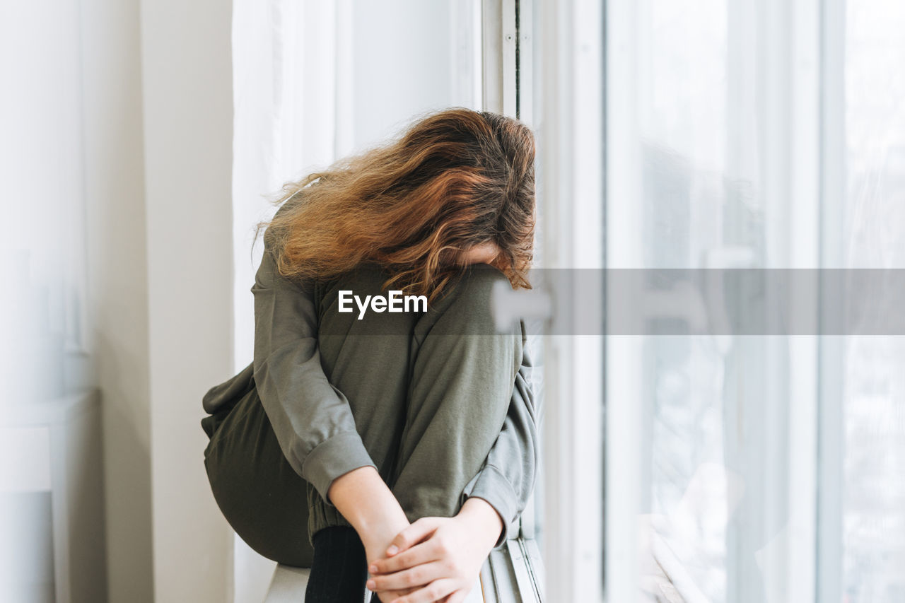 Beautiful sad unhappy teenager girl with curly hair in close pose sitting on window sill