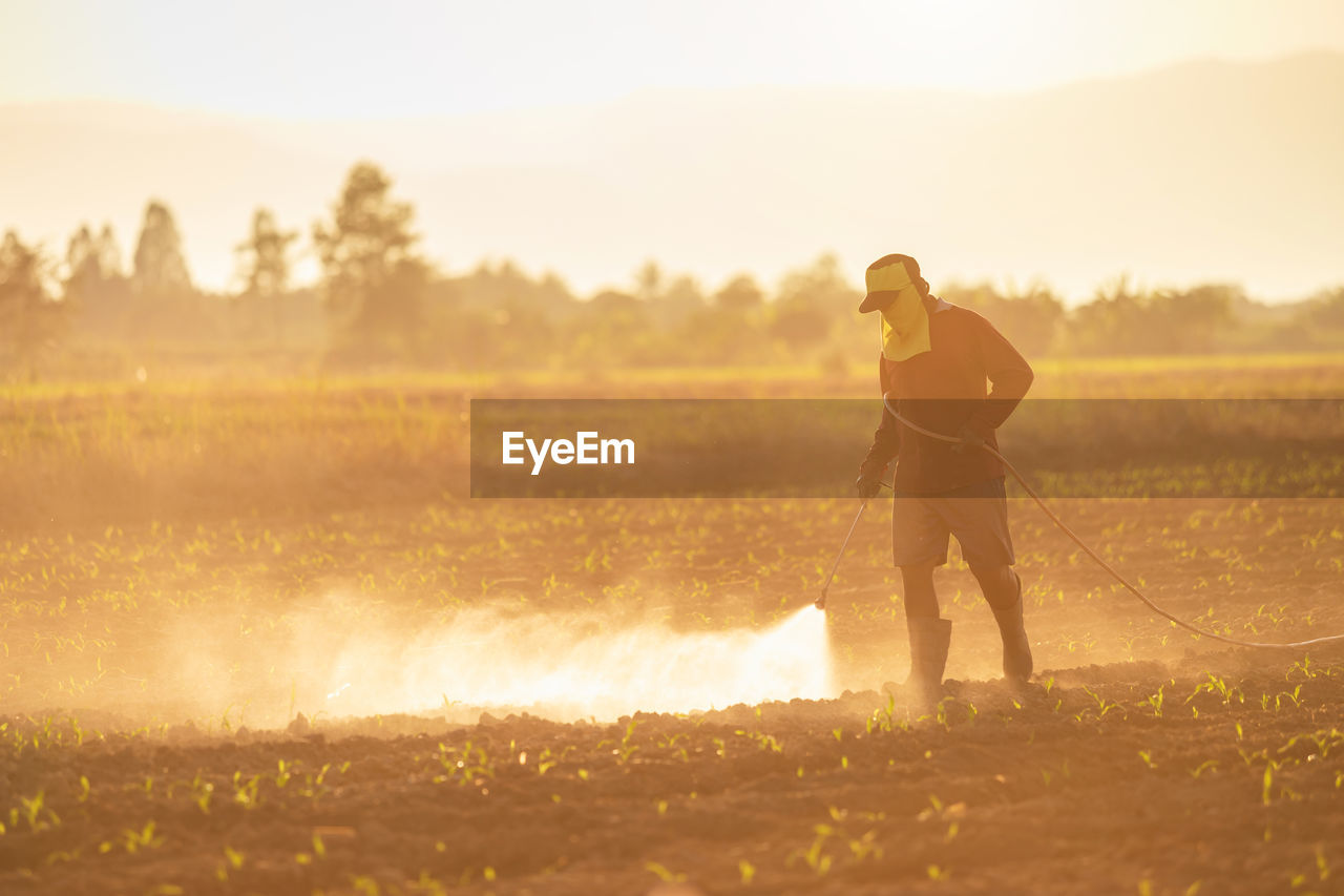 Full length of man spraying pesticides on field against sky during sunset