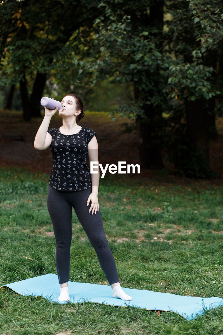 The relaxed girl is doing yoga in the park on carpet and is drinking w