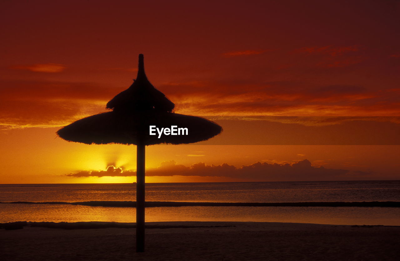 Silhouette thatched roof on beach against orange sky
