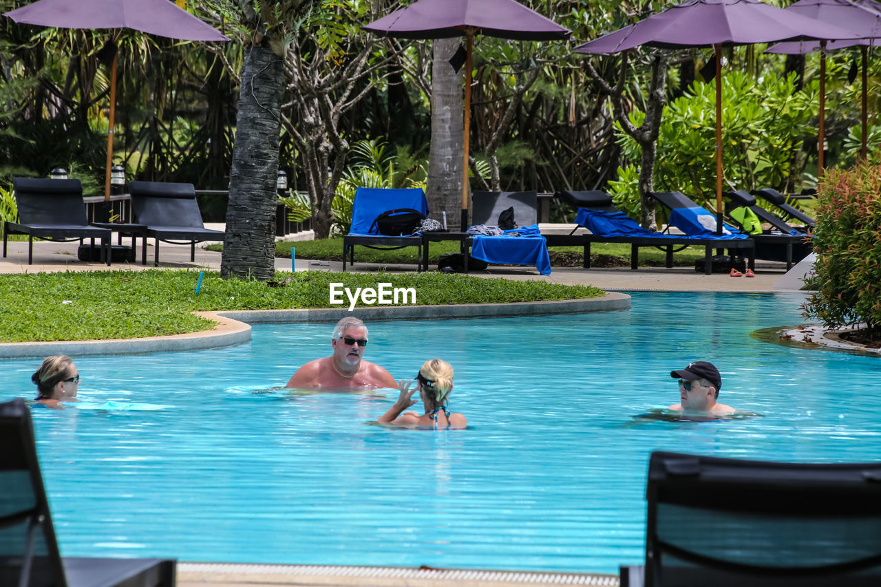 water, swimming pool, nature, swimming, adult, umbrella, relaxation, men, group of people, trip, leisure activity, women, vacation, holiday, enjoyment, lifestyles, parasol, tree, plant, lounge chair, tourist resort, wet, summer, resort, day, poolside, sunshade, chair, swimwear, fun, resort town, togetherness, outdoors, female, travel destinations, backyard, sitting, food and drink, sunlight, full length, small group of people, tropical climate, wealth