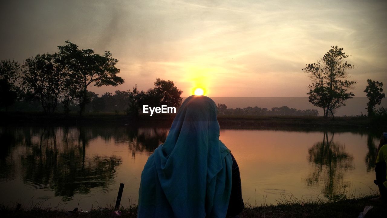 Rear view of woman at lakeshore against sky during sunset