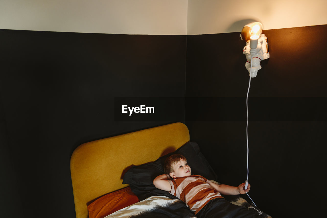 Boy turning on astronaut shape light lying on bed