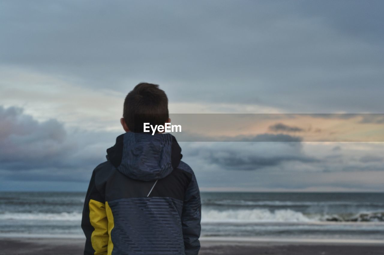 Rear view of boy standing at beach against cloudy sky