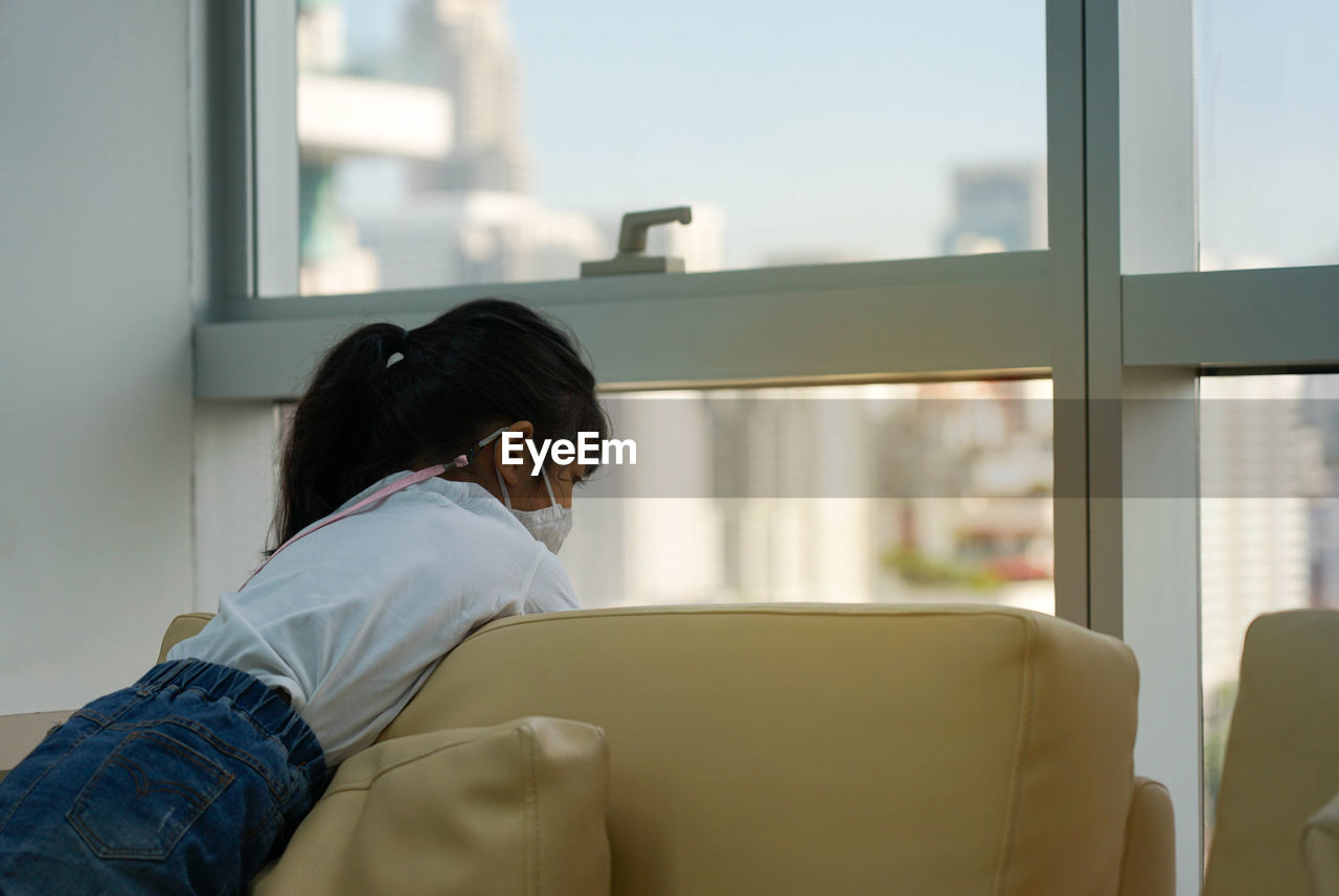 Asian cute kid stay at sofa at home she using fabric mask quarantine at home looking to the outside