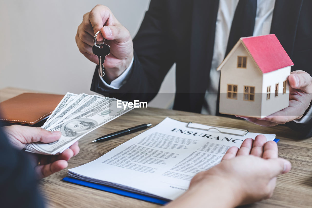 Midsection of real estate agent giving house key to customer in exchange of paper currency over table