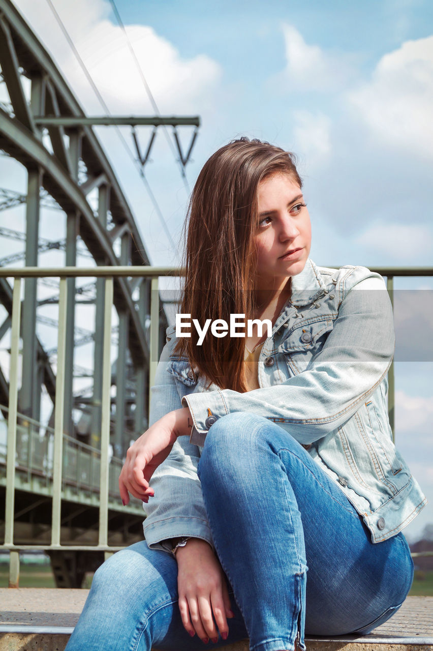 Beautiful young woman looking away while sitting against sky