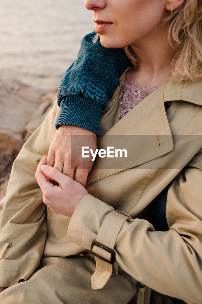 Crop man embracing blond girlfriend in outerwear and looking away while sitting on stony coast of rippling sea on cloudy day in aviles, spain