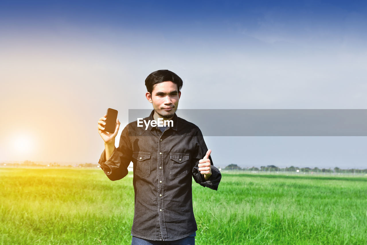 Portrait of man holding mobile phone while standing on grassy field against sky