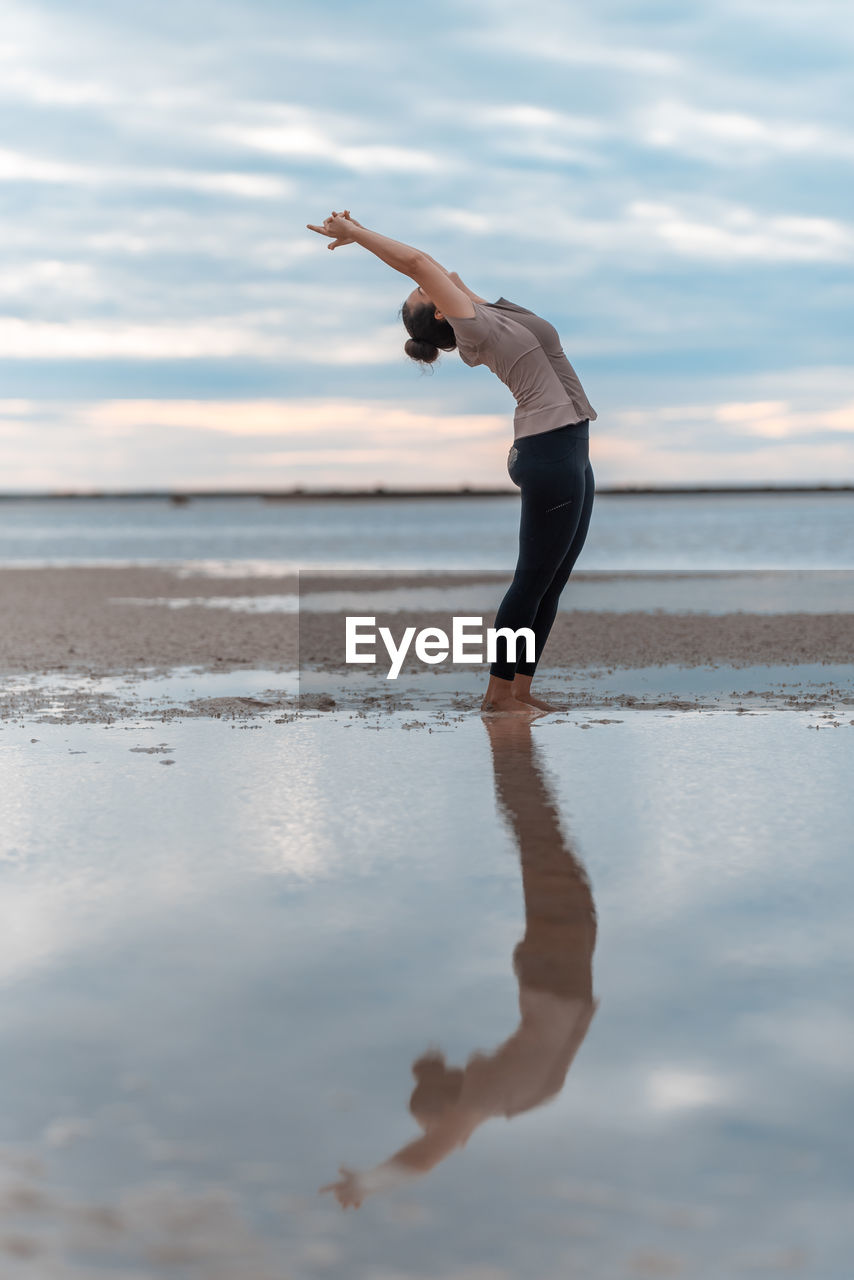 water, one person, sky, sea, adult, exercising, beach, nature, reflection, lifestyles, physical fitness, cloud, land, full length, sports, yoga, wellbeing, flexibility, balance, side view, stretching, vitality, leisure activity, tranquility, day, barefoot, beauty in nature, young adult, women, sand, relaxation, clothing, horizon over water, motion, standing, outdoors, arm, limb, tranquil scene, men, spirituality, female, human limb, holiday