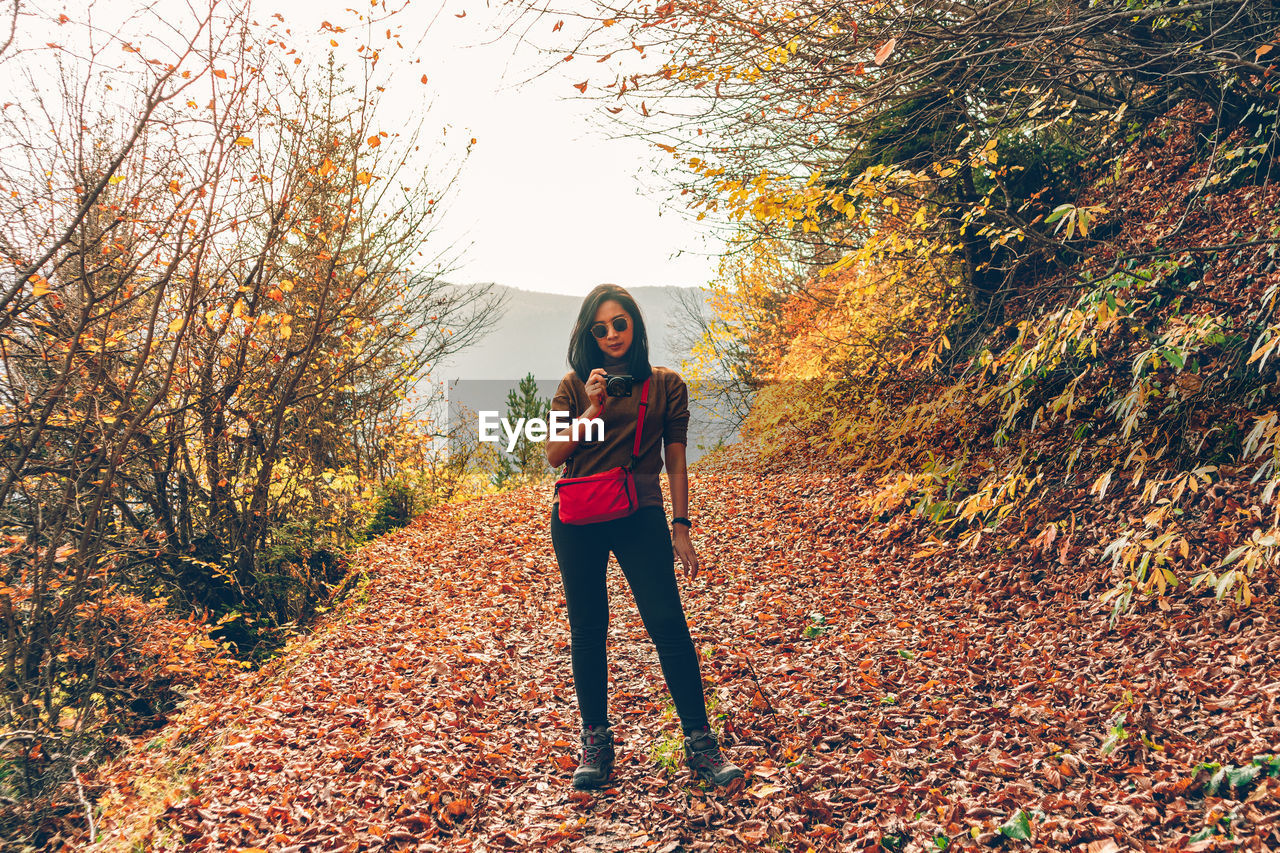 Full length of woman photographing while standing in forest during autumn
