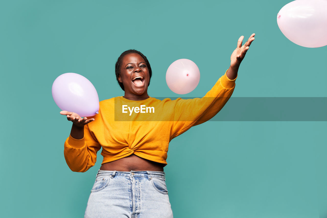 Excited african american female in jeans and yellow top standing near falling balloons against blue background in studio