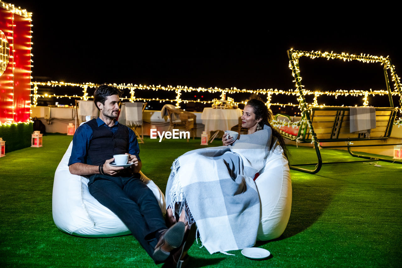 Young couple having coffee while relaxing on illuminated terrace at night