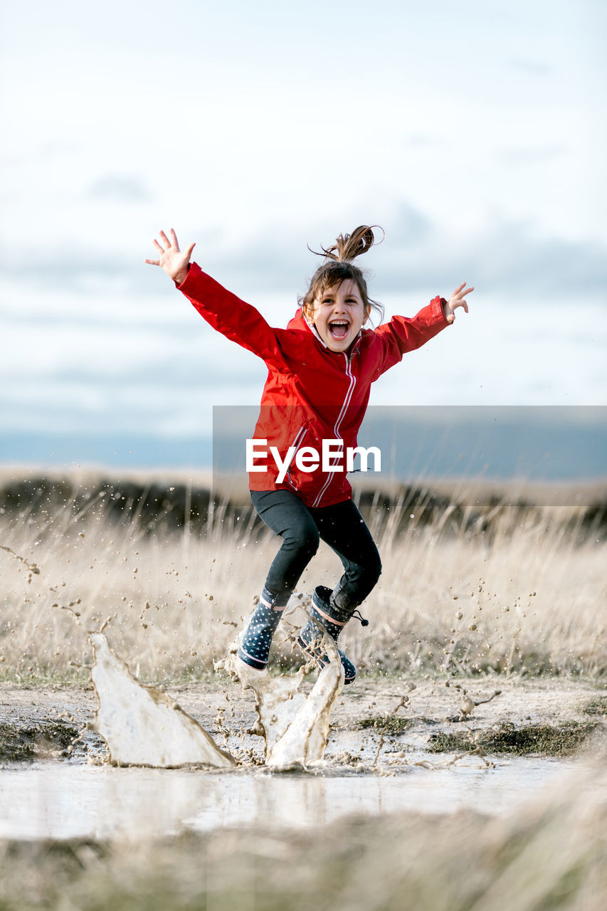 Carefree kid in raincoats and rubber boots splashing water in puddle while having fun and laughing on sunny day in countryside