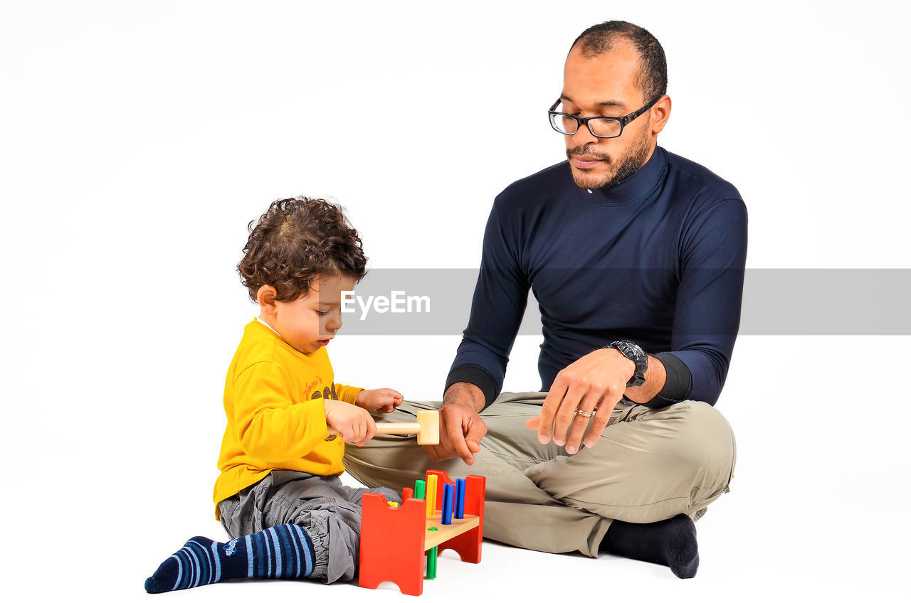 Father teaching son peg toy game while sitting against white background