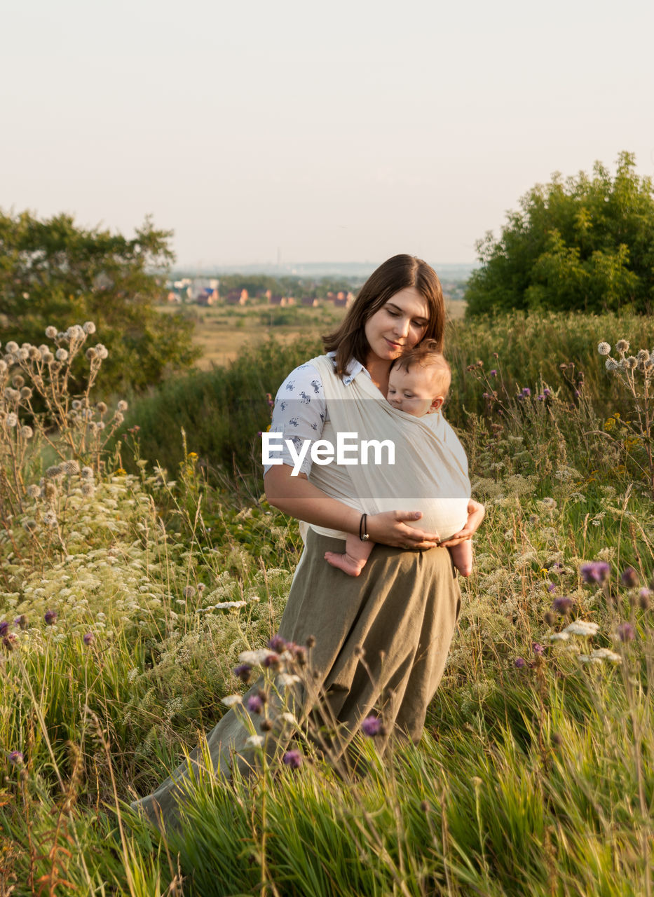 Woman with baby carrying in fabric while standing on field against sky