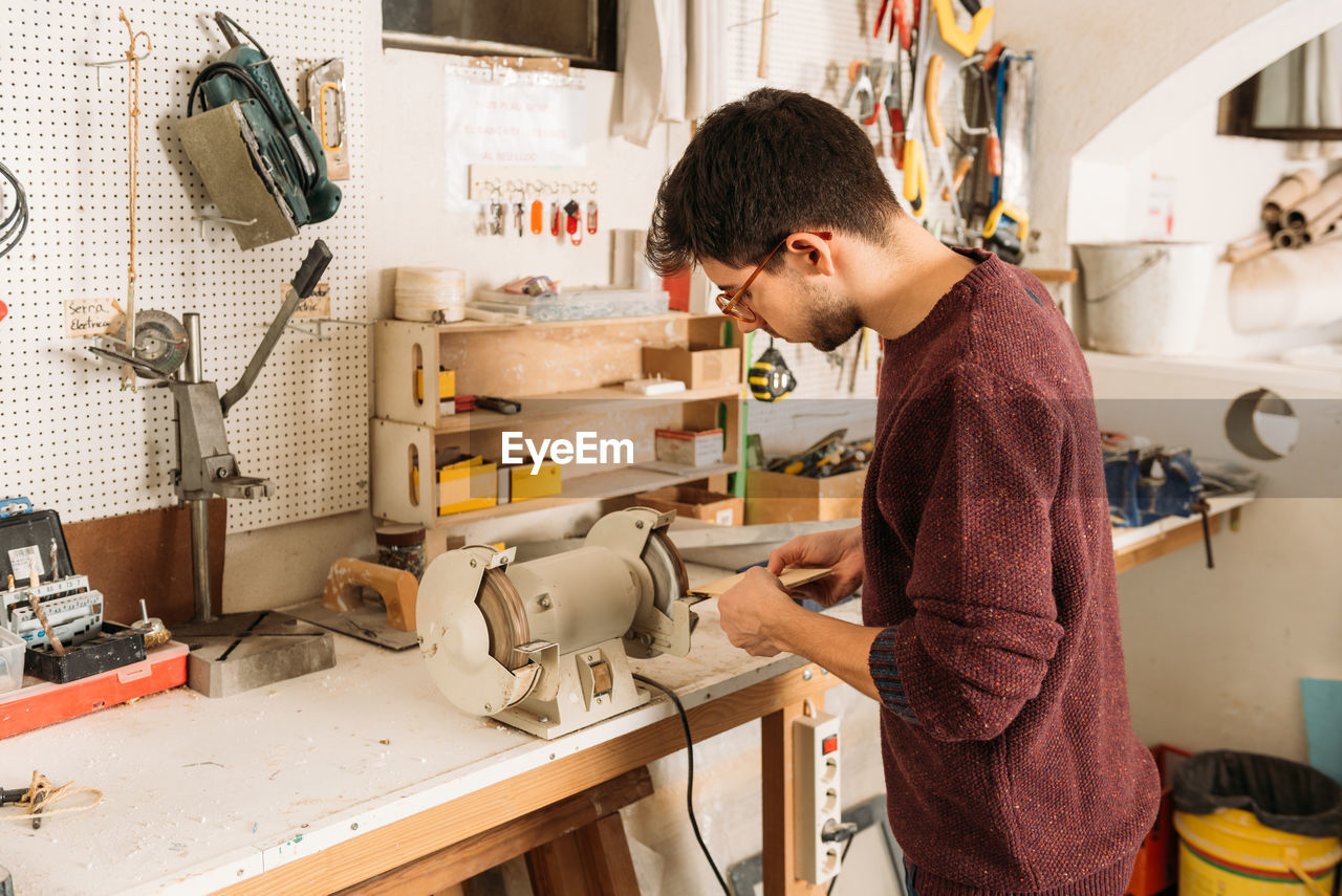 Side view of male carpenter using grinding machine for polishing wooden piece while working in bright workshop