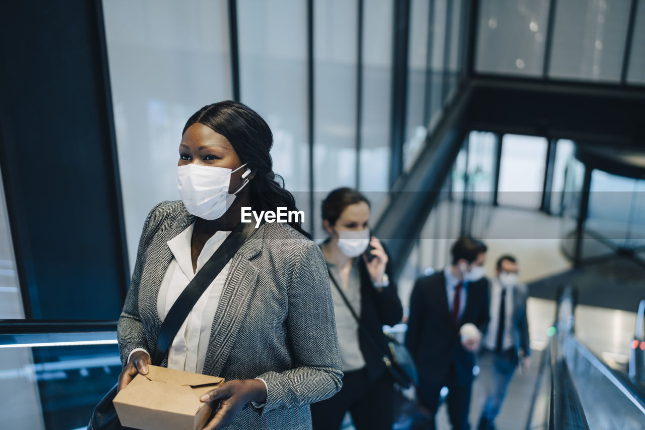 Female entrepreneur with box container standing on escalator during covid-19