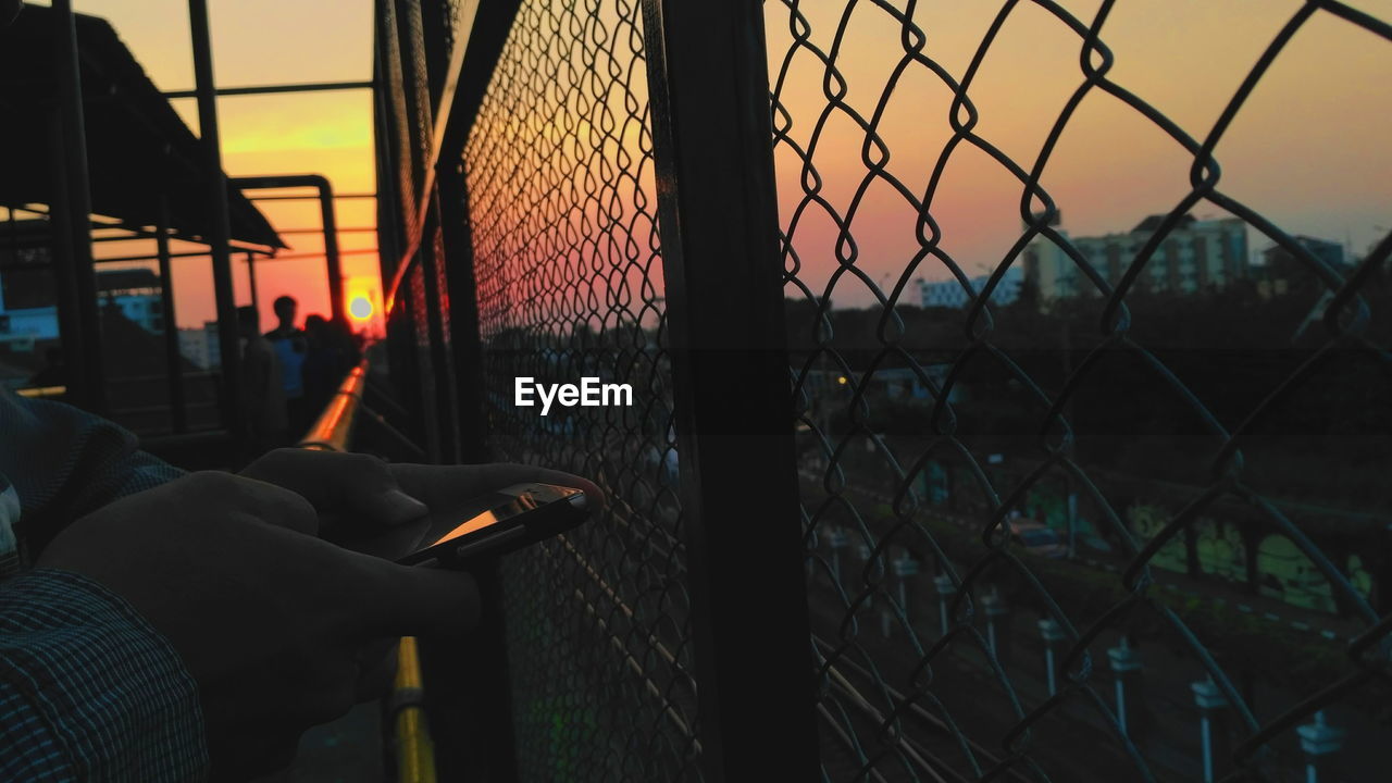 Close-up of cropped hands holding on chainlink fence against sunset sky