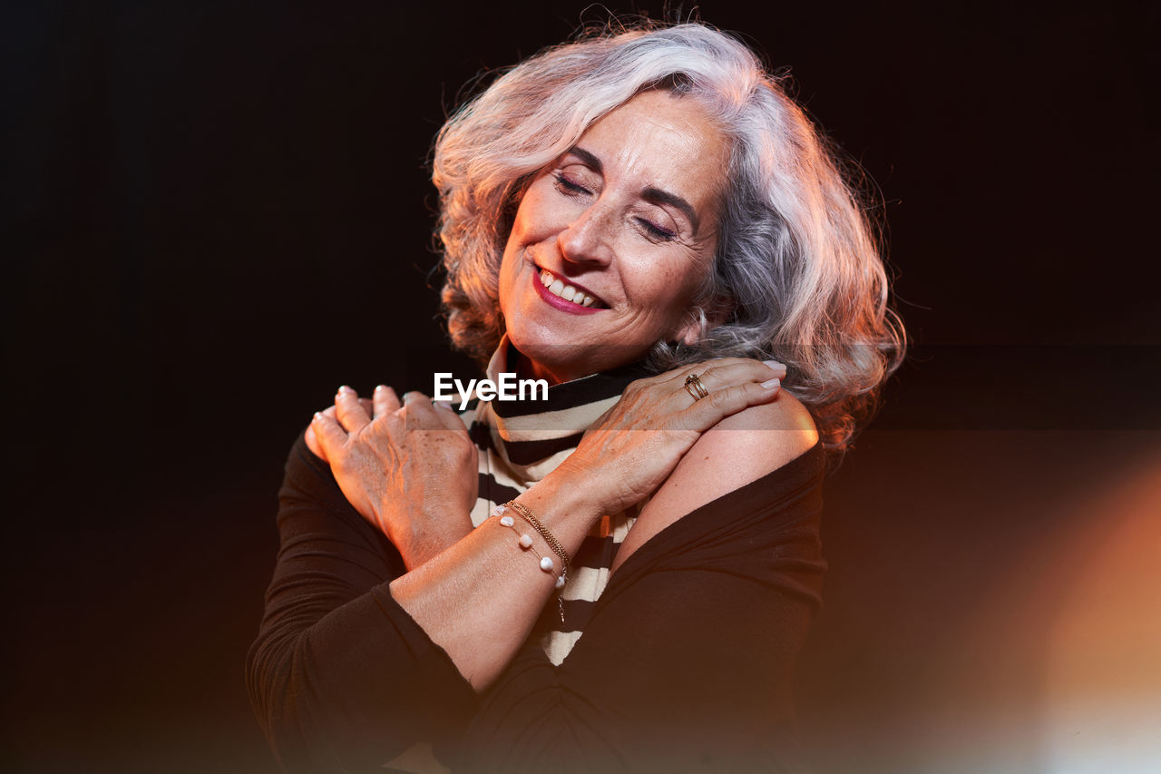 Happy senior female in stylish clothes with gray hair smiling with closed eyes and touching shoulders with crossed arms against black background