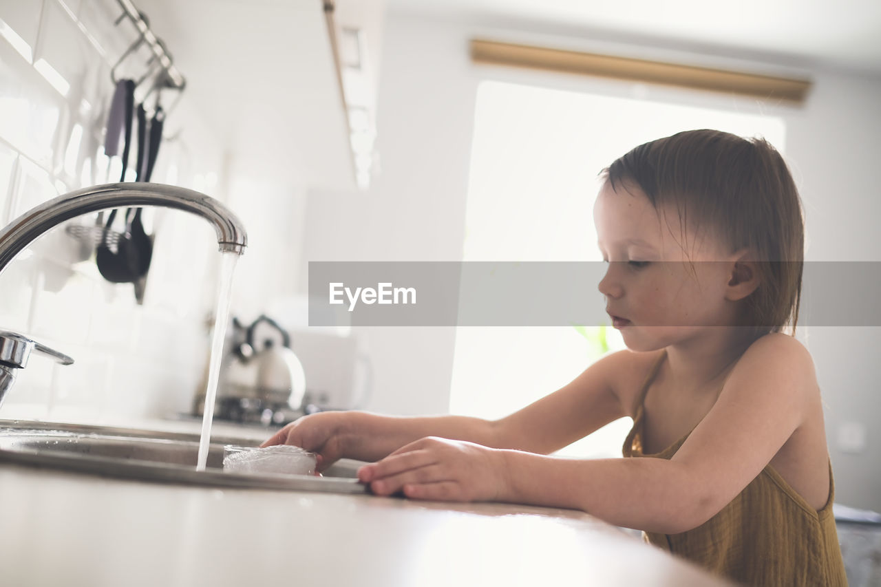 European child of 4 years old washes glass in sink on his own, bright kitchen in real interior, 