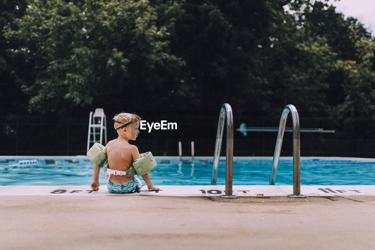 Rear view of shirtless boy wearing water wings sitting at poolside