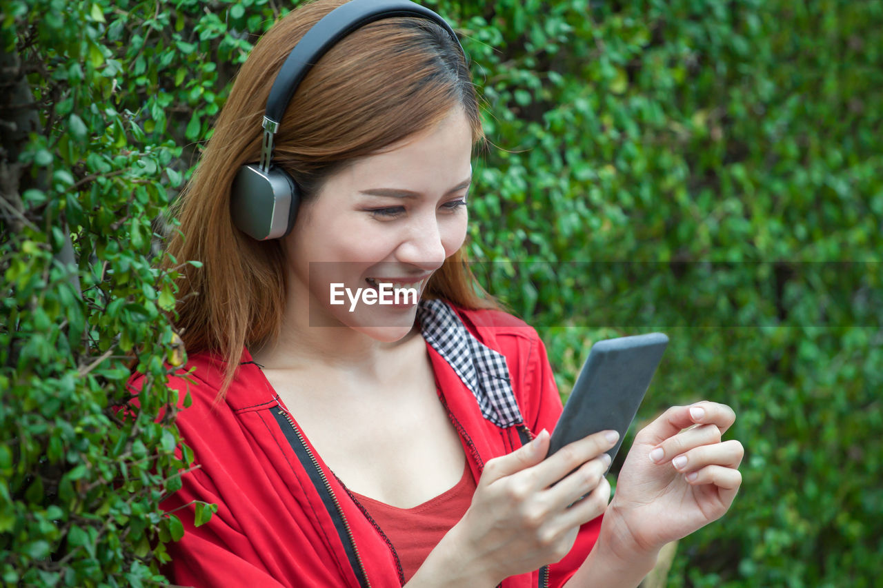 Smiling beautiful woman listening to music while leaning on hedge in garden