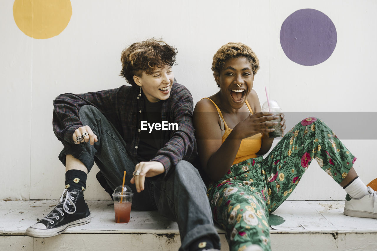 Cheerful friends sitting with drinks on retaining wall