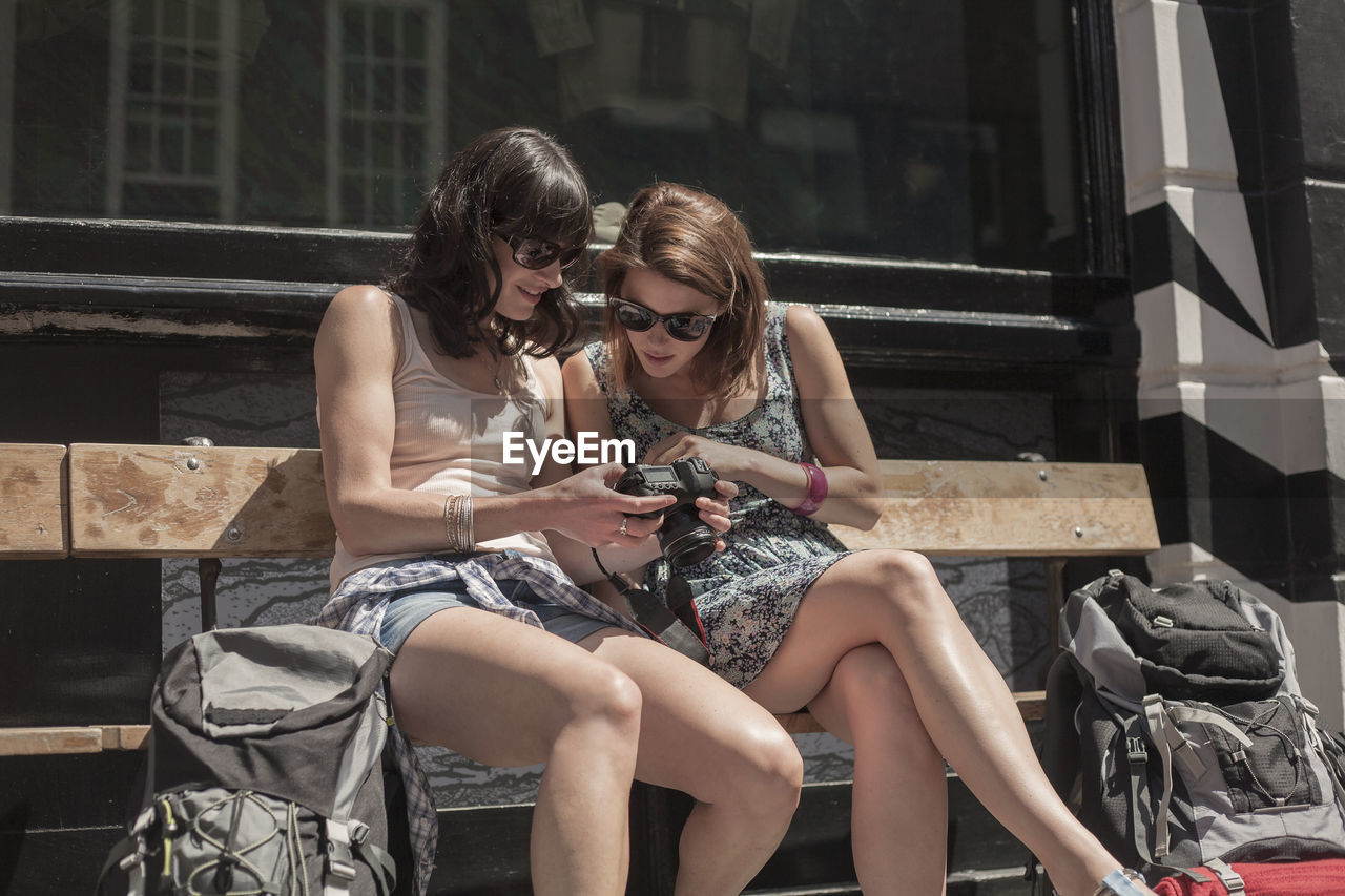 Curious woman looking at camera held by female friend while sitting on bench in city