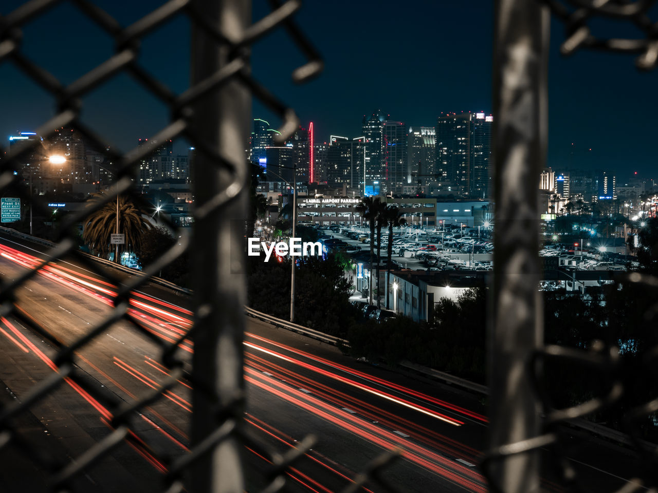 High angle view of light trails on road in city seen through broken chainlink fence at night