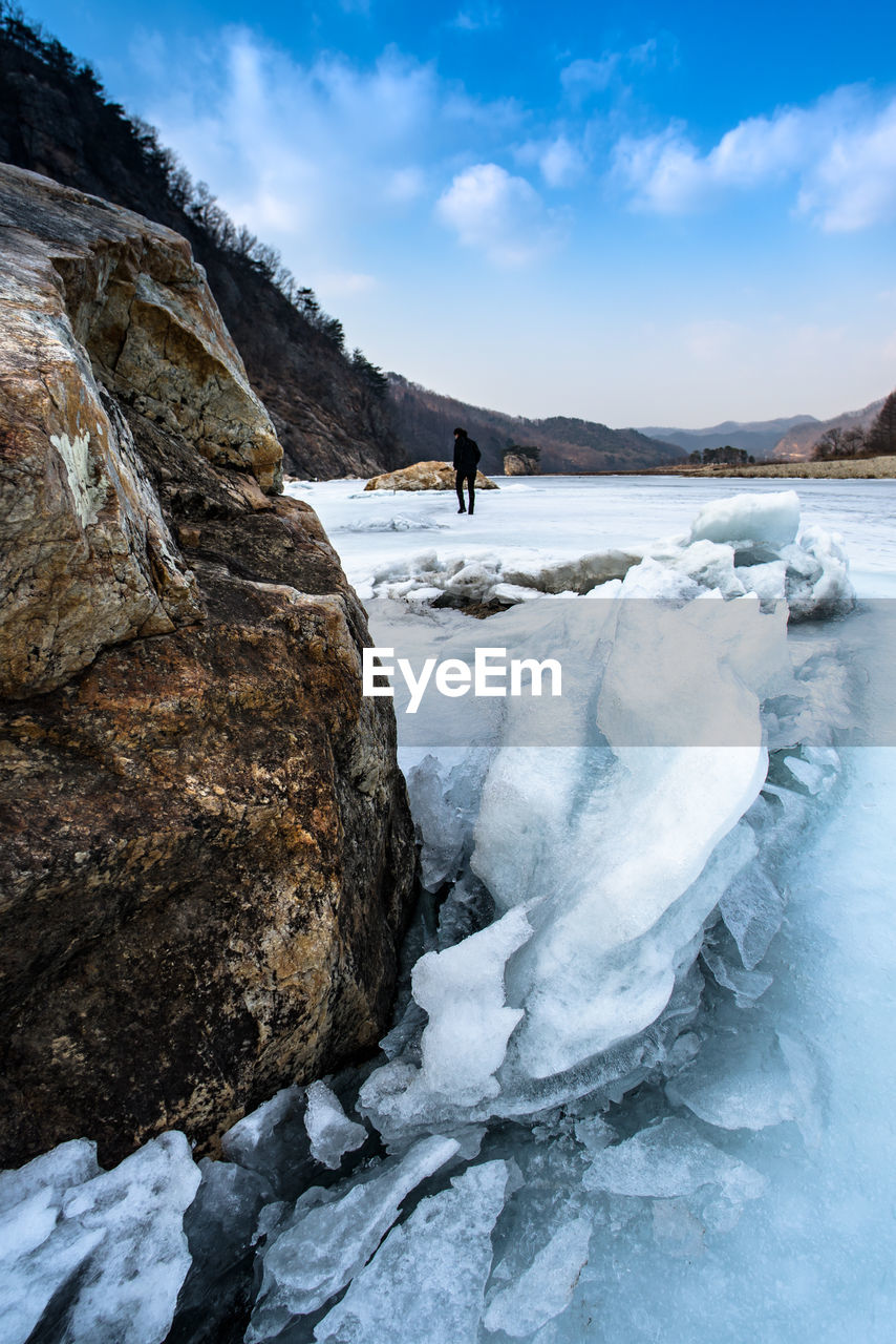 Woman walking by frozen river amidst mountains against cloudy sky