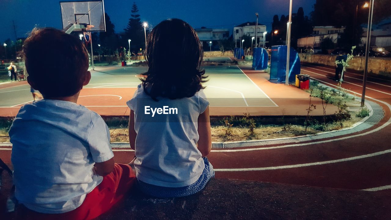 Rear view of kids sitting on retaining wall looking at illuminated basketball court