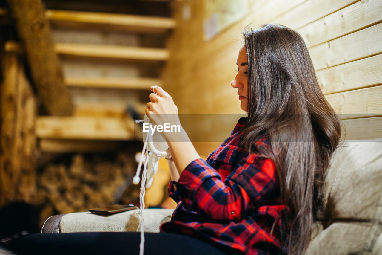 Side view of mid adult woman knitting while sitting on sofa at home