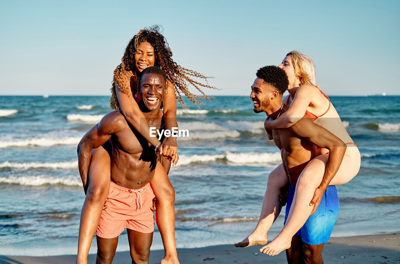 Happy multiracial women climbing on backs of black boyfriends while playing in foamy sea waves on summer day