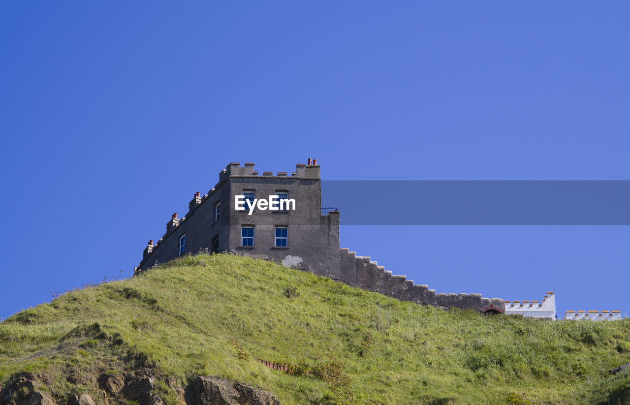 architecture, sky, built structure, building exterior, blue, tower, history, clear sky, the past, building, hill, nature, travel destinations, fort, castle, land, travel, no people, plant, low angle view, tourism, terrain, copy space, fortification, fortified wall, outdoors, cliff, day, coast, scenics - nature, ancient, sunny, landscape, landmark, sea, mountain range, grass, house, beauty in nature, old, wall