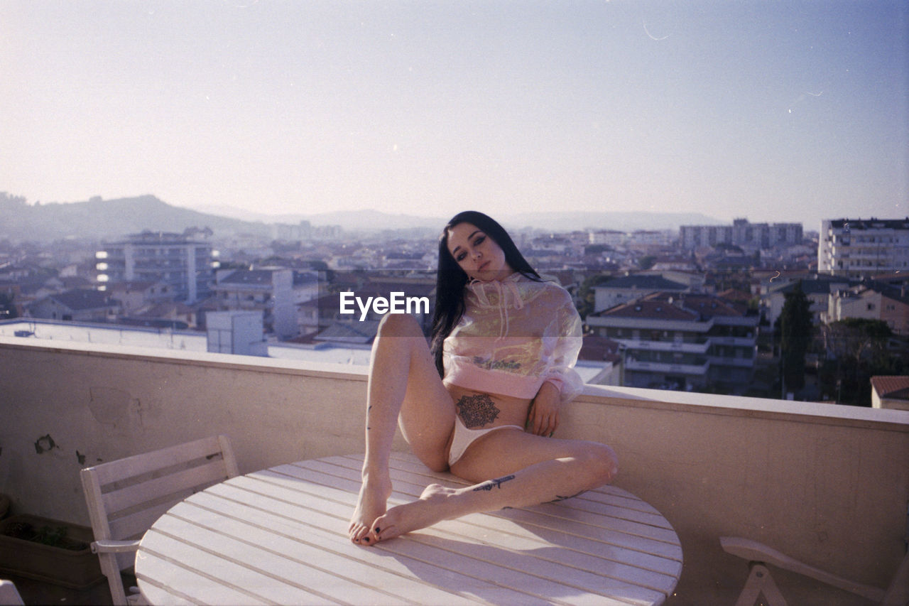 PORTRAIT OF WOMAN SITTING ON BALCONY AGAINST CITYSCAPE