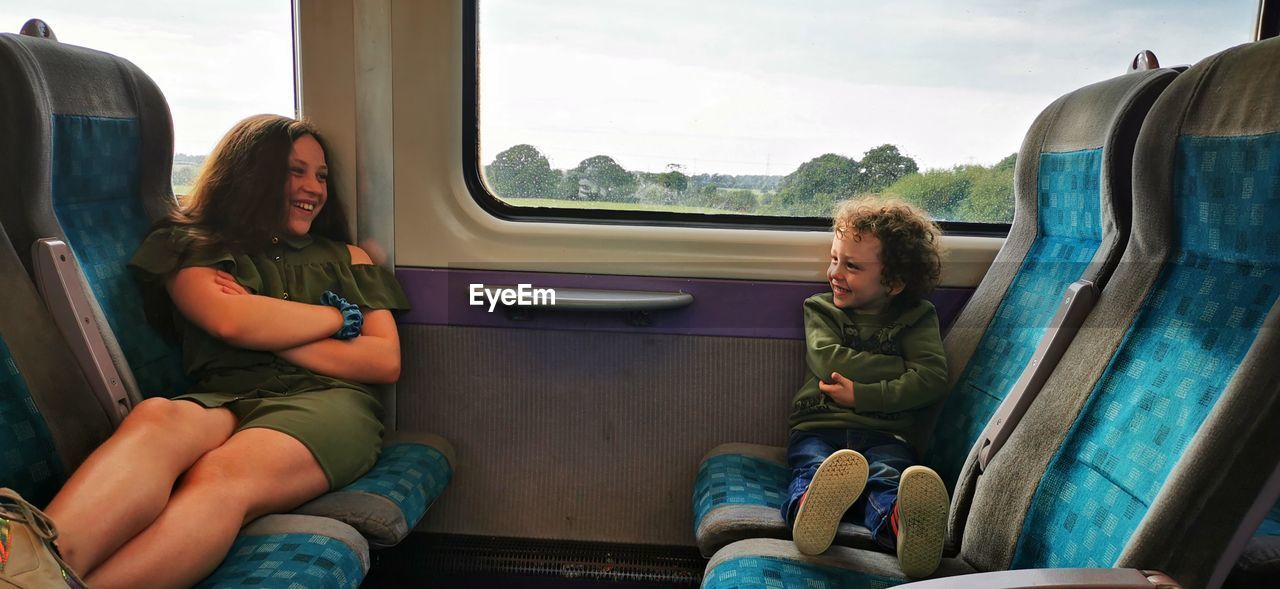 Smiling siblings with arms crossed sitting on chairs against window in train