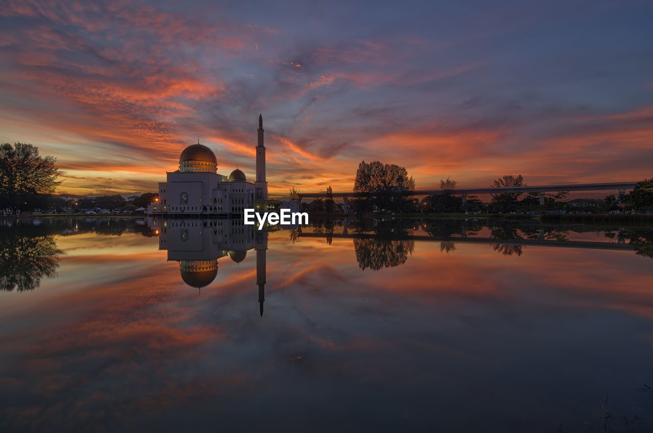 Reflection of trees and mosque on lake against sky during sunset
