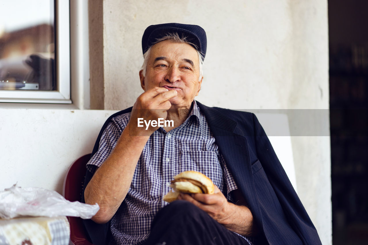 Senior man eating burger while sitting against wall