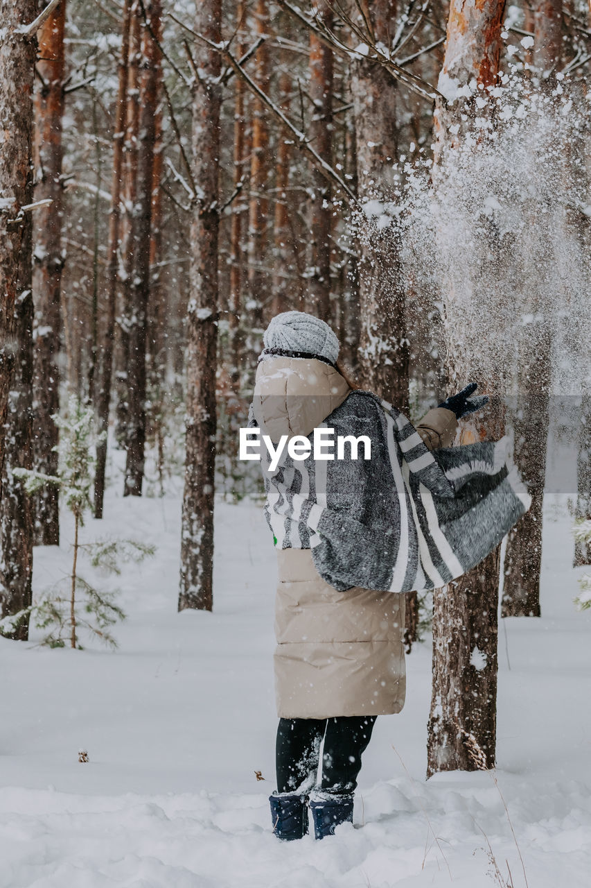 A girl in a striped scarf stands in a winter forest and throws snow in the air.