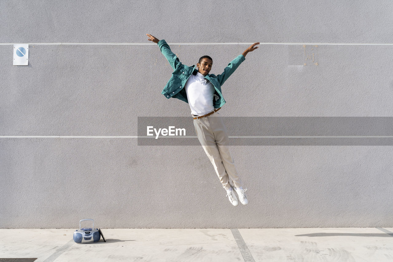 Young man with radio on footpath jumping against gray wall