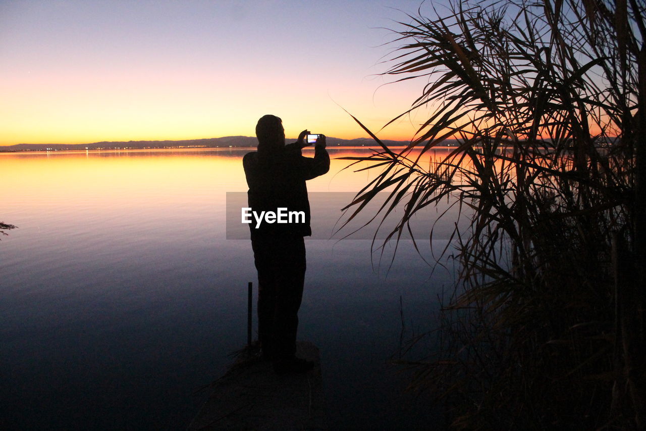 Silhouette man photographing by lake against sky during sunset